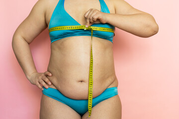 Measure female breast with tape and determine number closeup, folds on stomach, cellulite. Naked overweight plus size girl on pink background in blue underwear. Concept of dieting and body control.
