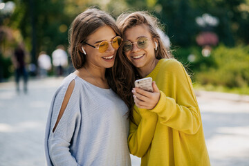 Fototapeta premium Girls outside in bright clothes and sunglasses in headphones, holding smartphone. Video call, online time with friends, outdoor time, warm sunny day in city