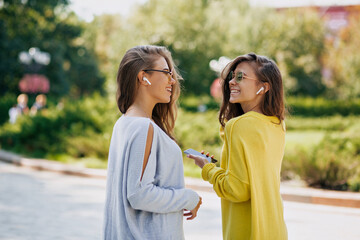 Pretty modern women with long wavy hairstyle in bright clothes are talking and listening music in sunny warm day on city background