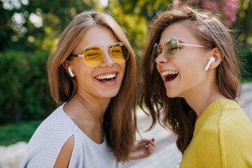 Cheerful smiling woman with widely smile laughing with eyes closed while listening music with sister. Outdoor photo of two emotional female friends.