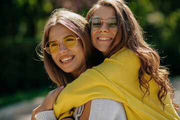 Pretty charming lady with long wavy hair in bright sweater is hugging her sister and laughing. Two stylish girls smiling in sunlight and wearing round sunglasses and bright pullovers