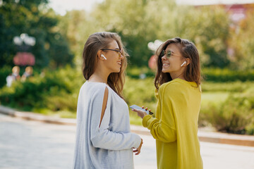 Two young ladies wearing yellow and blue sweaters are walking outside in sunny city and listening music in headphones 