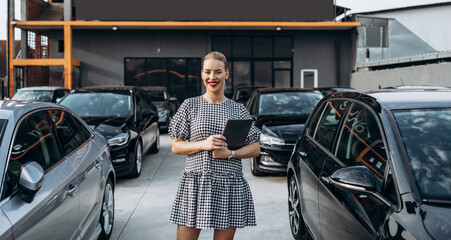 Beautiful young female car dealer standing in front of parked cars. Used car dealership concept.