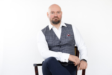 Portrait of confident middle-aged businessman in grey vest, blue jeans, white shirt, sitting on chair with legs crossed.