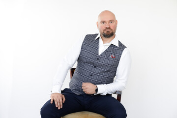 Portrait of bald middle-aged businessman in grey vest, blue jeans, white shirt sitting, leaning...