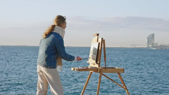 Art hobby. Young inspired talented woman creating her masterpiece on seashore, drawing on wooden easel on sea bank
