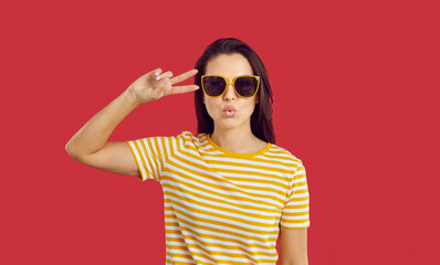 Fototapeta premium Studio shot of good looking young woman in trendy sunglasses. Happy confident attractive brunette lady in striped T shirt and cool glasses sending you a kiss and showing peace, love and victory sign