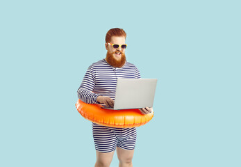 Work and vacation. Funny excited chubby man on pastel turquoise background working remotely with laptop during summer vacation. Red-bearded man in striped leotard with inflatable circle at waist.