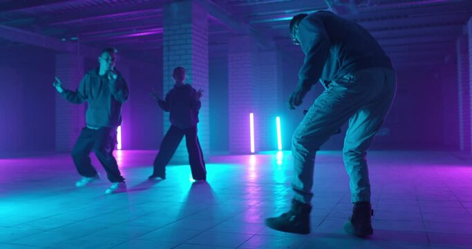 Backstage of video shooting. Cameraman filming hip-hop dancers in neon light. Operator moves with image stabilizer in hands at dark studio. Shooting a clip