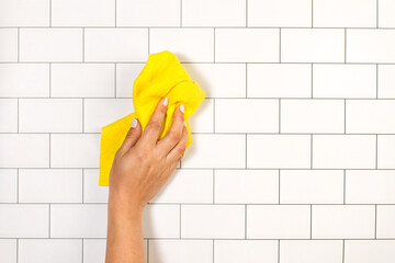 Mano de mujer limpiando con un paño amarillo una pared con azulejos. Vista de frente y de cerca. Copy space - obrazy, fototapety, plakaty