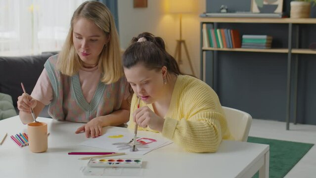 Young woman and girl with Down syndrome painting picture on paper with watercolor paints while sitting at desk at home