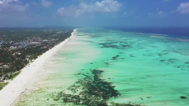 High forward-moving drone aerial view of Paje village, the beach and its blue lagoon, and the coral reef, with people kitesurfing
