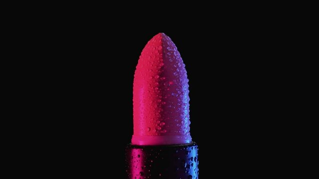 A tube of red female lipstick in raindrops rotates on a black background. Macro shooting.