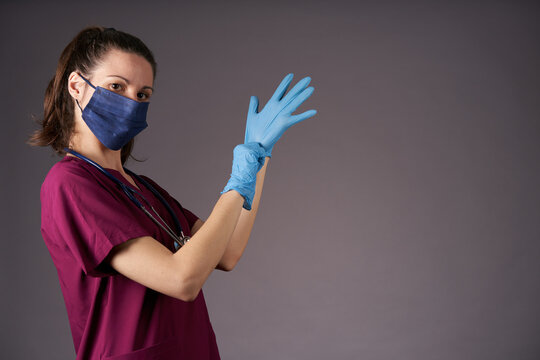 Nurse In Purple Uniform Mask And Stethoscope Putting On Latex Medical Gloves Looking At Camera On Gray Background