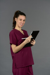 female doctor in purple uniform filling out report and looking at camera on digital tablet on gray background