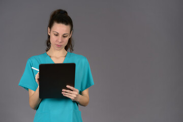 Obraz premium smiling female surgeon in aquamarine scrubs filling out pre-surgery report on digital tablet on gray background