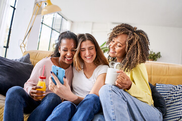 Three cheerful happy woman watching video using mobile smart phone at home together sitting at sofa...