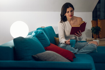 Woman reading a book and drinking tea while relaxing on a sofa at home