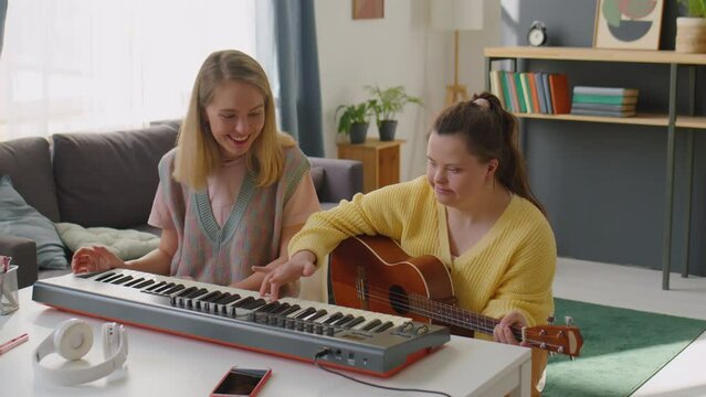 Female teacher smiling and looking at girl with Down syndrome sitting with guitar and playing synthesizer during music lesson at home