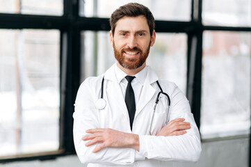 Caucasian handsome male doctor, therapist, surgeon, cardiologist in a medical uniform and stethoscope, standing in a hospital, looking to the camera with arms crossed, smiling friendly