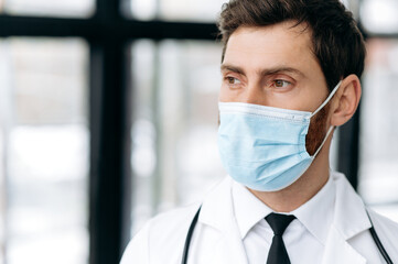 Close-up of Caucasian pensive male doctor, therapist, surgeon, cardiologist in a medical uniform, protective mask and with stethoscope, standing in a hospital, looking to the side with, thinking