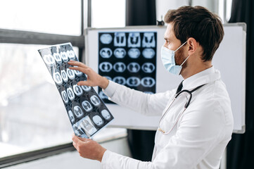 Fototapeta na wymiar Smart professional Caucasian doctor in a medical uniform, protective mask, a pulmonologist, a therapist, stands in hospital, examines an x-ray of his patient's lungs,thinks about prescribing treatment