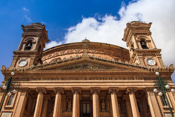 Fototapeta premium Malta island Mosta Rotunda, the Sanctuary Basilica of the Assumption of Our Lady - Santwarju Bazilika ta Santa Marija, 17th century Catholic church facade against blue sky with clouds.