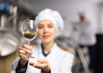 Successful smiling female brewer in white uniform showing natural granulated hops while standing in brewery. Selective focus on glass with pellets. Concept of high quality raw materials..