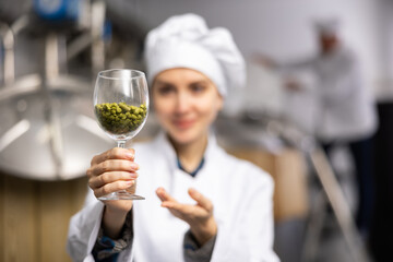 Successful smiling female brewer in white uniform showing natural granulated hops while standing in brewery. Selective focus on glass with pellets. Concept of high quality raw materials..