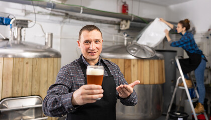 Portrait of brewer who is standing with glass of beer on his workplace in the brew-house
