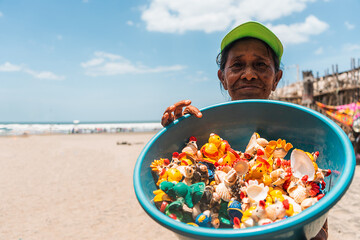 Elderly Latina street vendor on the beach displaying a bowl full of handicrafts made from painted...
