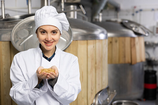 Portrait of positive woman brewmaster holding bunch of malt seeds in beer factory. - Powered by Adobe