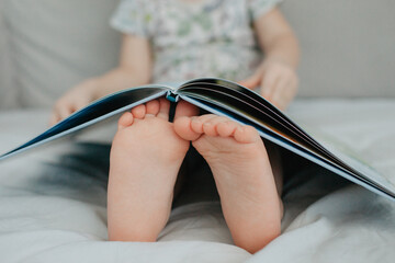 little girl holding bare feet close up to camera and reading book . Blurred face on background