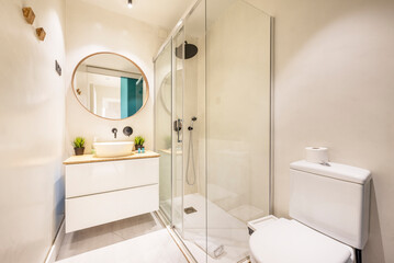 Washroom with shiny wooden hanging cabinet with circular mirror with wooden frame and shower cabin with glass partitions
