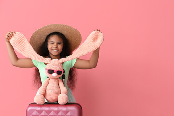 Cute little African-American girl with suitcase and bunny toy on pink background