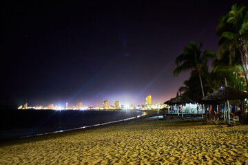 playa en la noche con palmeras en primer plano una ciudad con luces brillantes en el fondo y un...
