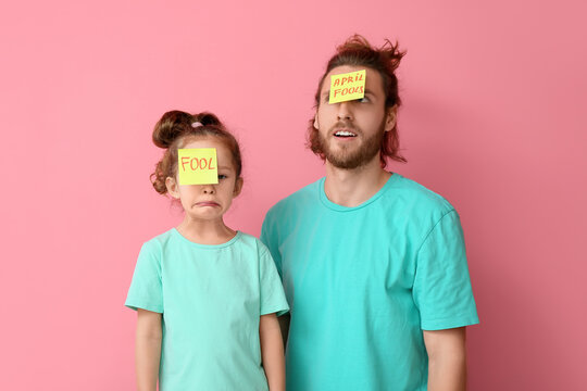 Funny man and her little daughter with sticky notes on their foreheads against pink background. April fools' day celebration