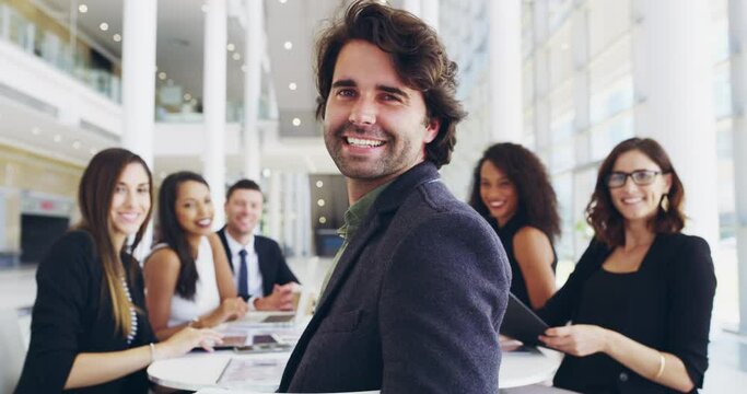 We always work as an empowered team. A young businessman smiling in an office during a meeting with his colleagues in the background. Diverse group of working professionals brainstorming together 
