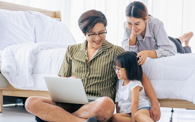 Obraz premium Asian Father, Mother, little daughter girl sitting in bedroom at home, using laptop, online studying, doing homework, watching movie, playing games, shopping together with happiness. Family Concept.