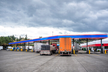 Fototapeta na wymiar Big rig semi trucks with different semi trailers and loads refills the semi trucks tanks at truck stop gas station to continue the cargo freights
