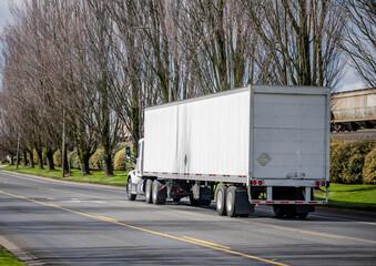 Big rig white day cab semi truck tractor transporting cargo in dry van semi trailer moving on the city street road with trees alley