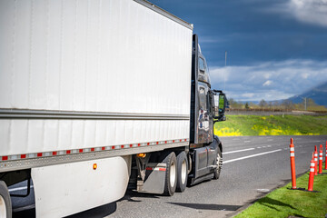 Black big rig bonnet semi truck transporting cargo in dry van semi trailer with aerodynamic skirt running on the local road with road work traffic cones