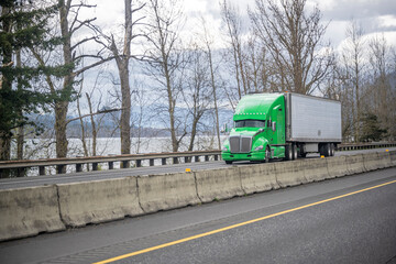 Bright green big rig semi truck transporting frozen cargo in refrigerator semi trailer running on the highway road along the river