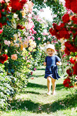 Portrait of little toddler girl in blossoming rose garden. Cute beautiful lovely child having fun with roses and flowers in a park on summer sunny day. Happy smiling baby.