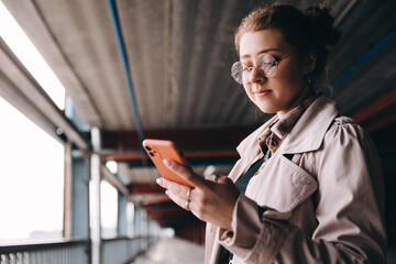 young caucasian female student, generation z, dressed in a trench coat in the parking lot uses a smartphone