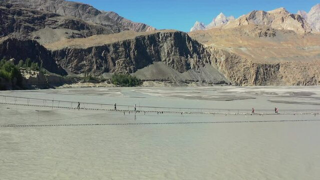 aerial drone flying backwards revealing the Passu Cones mountains in the distance as tourists cautiously cross the famous Hussaini Bridge in Hunza Pakistan as a fast paced gray river flows below.