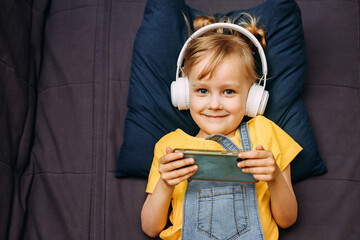 Happy boy with headphones sitting at home on the couch. The child is happy to learn at home online, listening to music. A new app for teaching children.