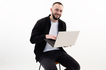 Young smiling bearded man with short dark hair wearing black jacket, jeans, white T-shirt, sitting on stool with laptop.