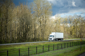 Obraz premium Long haul industrial big rig semi truck with dry van semi trailer transporting goods running on the divided highway road with bare trees on the side