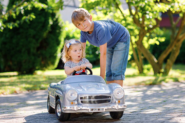 Two happy children playing with big old toy car in summer garden, outdoors. Boy driving car with little girl, cute sister inside. Laughing and smiling kids. Family, childhood, lifestyle concept.